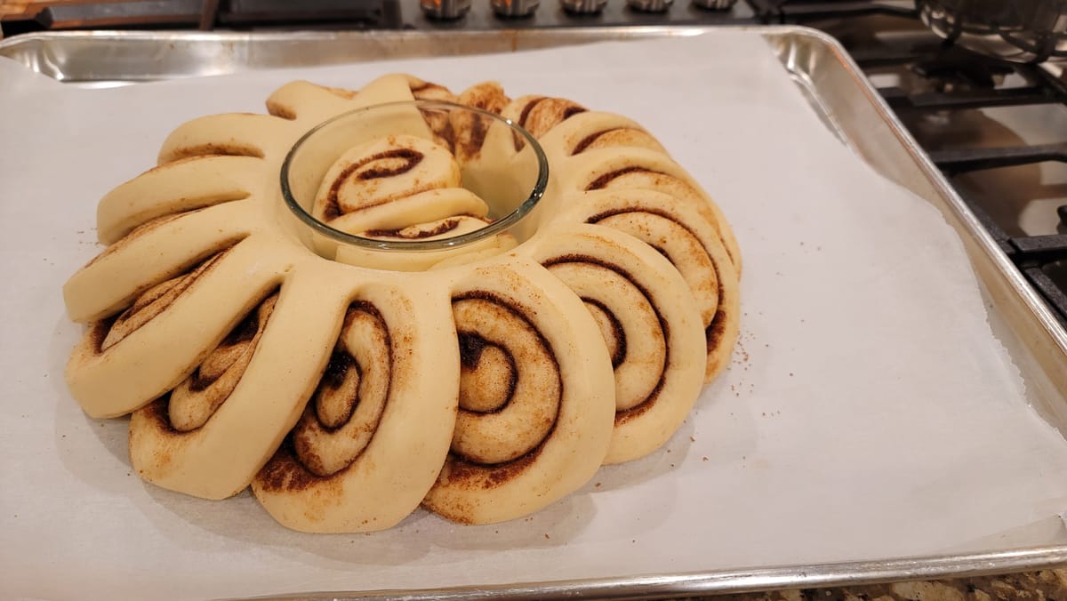 Golden-brown cinnamon bun wreath arranged in a circle on a white plate with white icing drizzle and cinnamon swirls visible