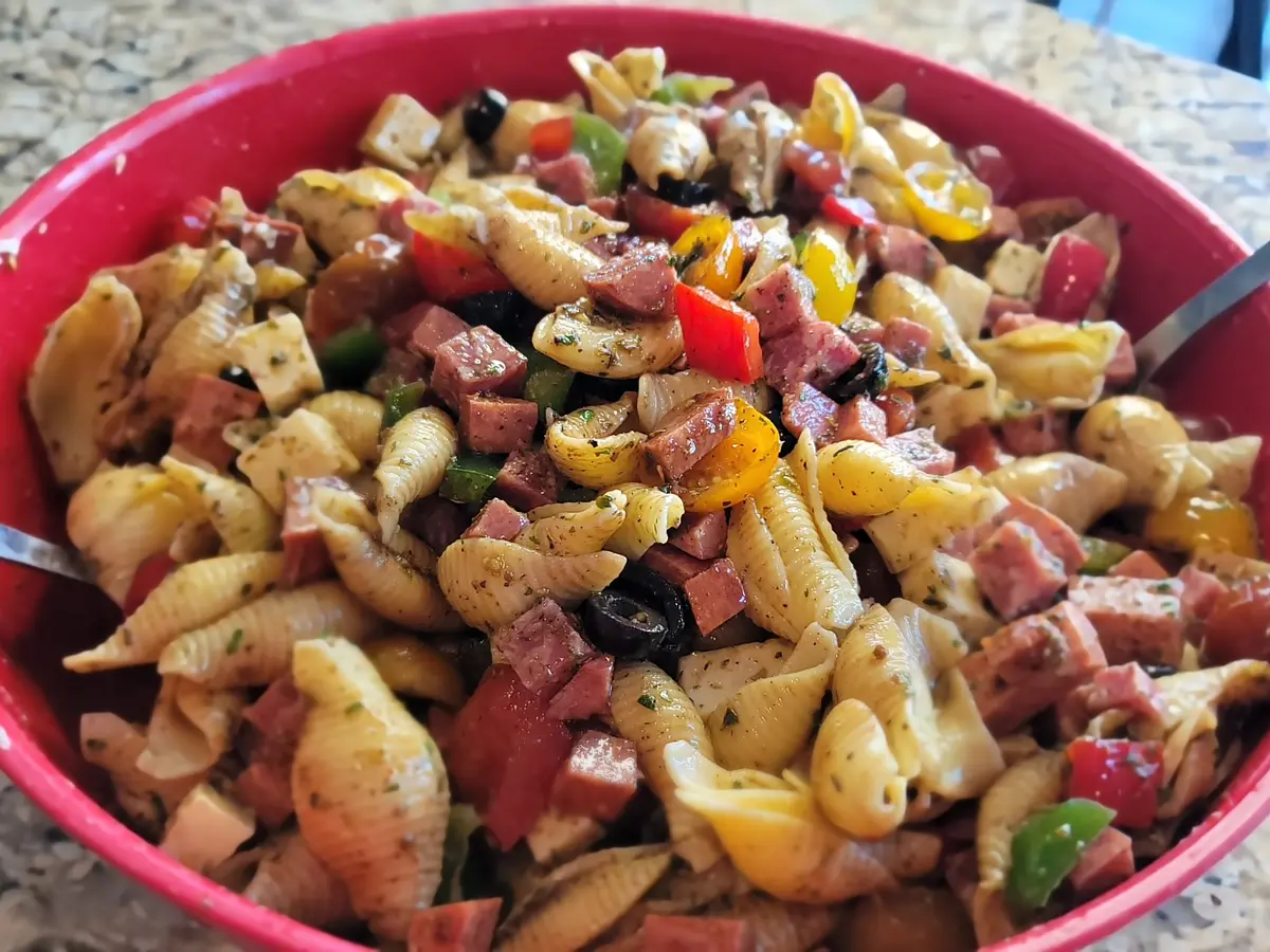 Colorful antipasto salad with shell pasta, diced salami, pepperoni, Asiago cheese, cherry tomatoes, bell peppers, and black olives in a serving bowl