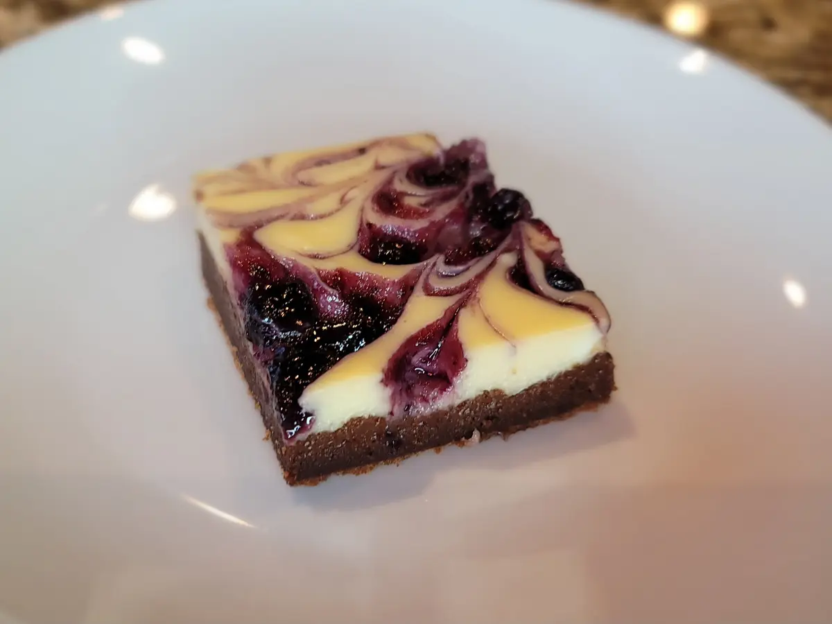 Golden-brown cheesecake brownie with vibrant raspberry swirl on a white plate, showing fudgy chocolate layers and creamy cheesecake texture