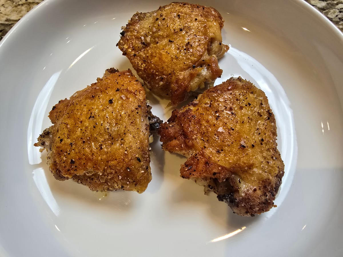 Golden crispy chicken thighs with bronzed skin on a white plate, showing the juicy meat inside