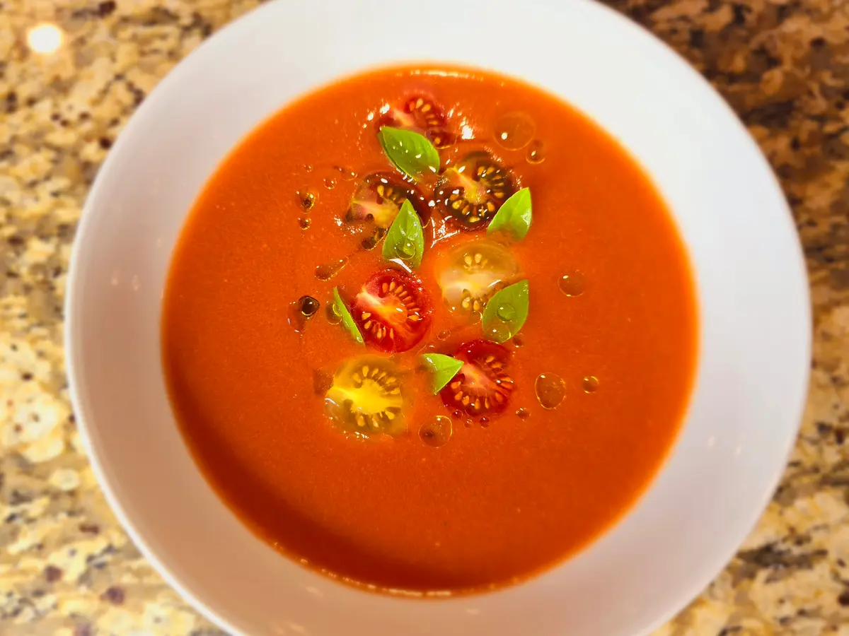 Bowl of vibrant red gazpacho soup garnished with diced vegetables and olive oil drizzle, fresh tomatoes and cucumber visible