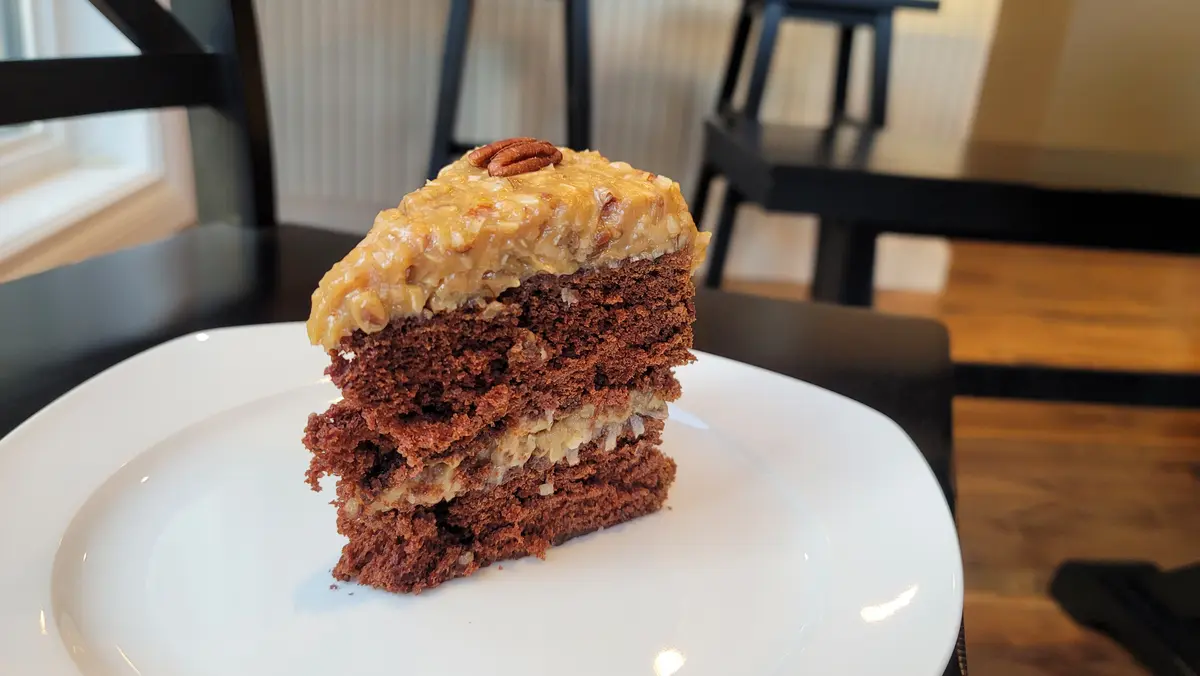Slice of German chocolate cake with two thick chocolate layers and gooey coconut-pecan frosting filling on a white plate