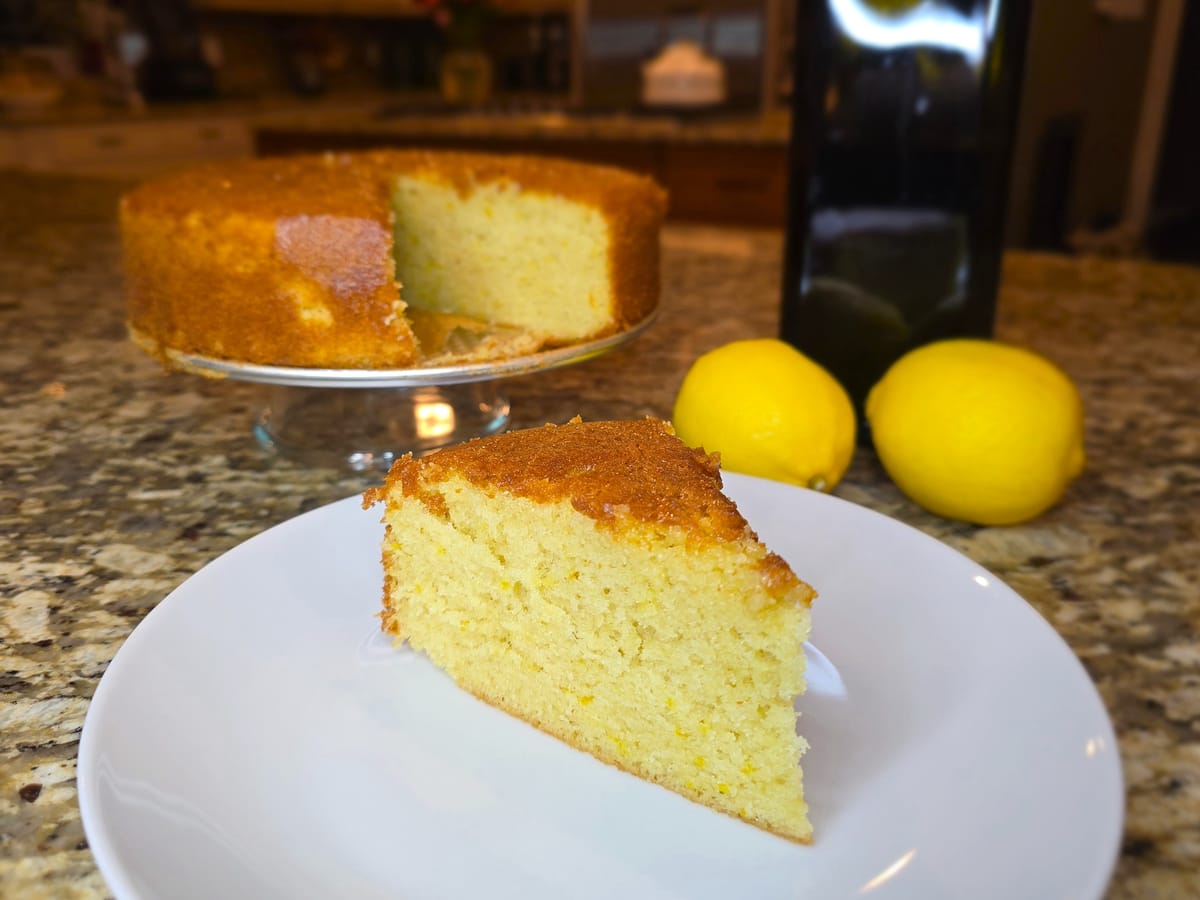 Slice of golden lemon olive oil cake on a white plate with fresh lemon zest and a fork