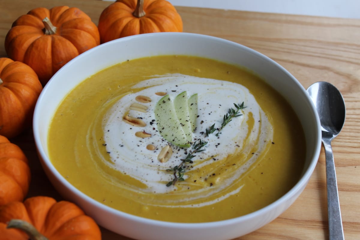 Creamy pumpkin soup in a white bowl topped with maple whipped cream and fresh thyme, garnished with autumn leaves