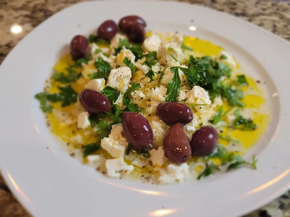 Zesty bread dip with crumbled feta, olives, and fresh parsley in a white bowl with olive oil drizzle