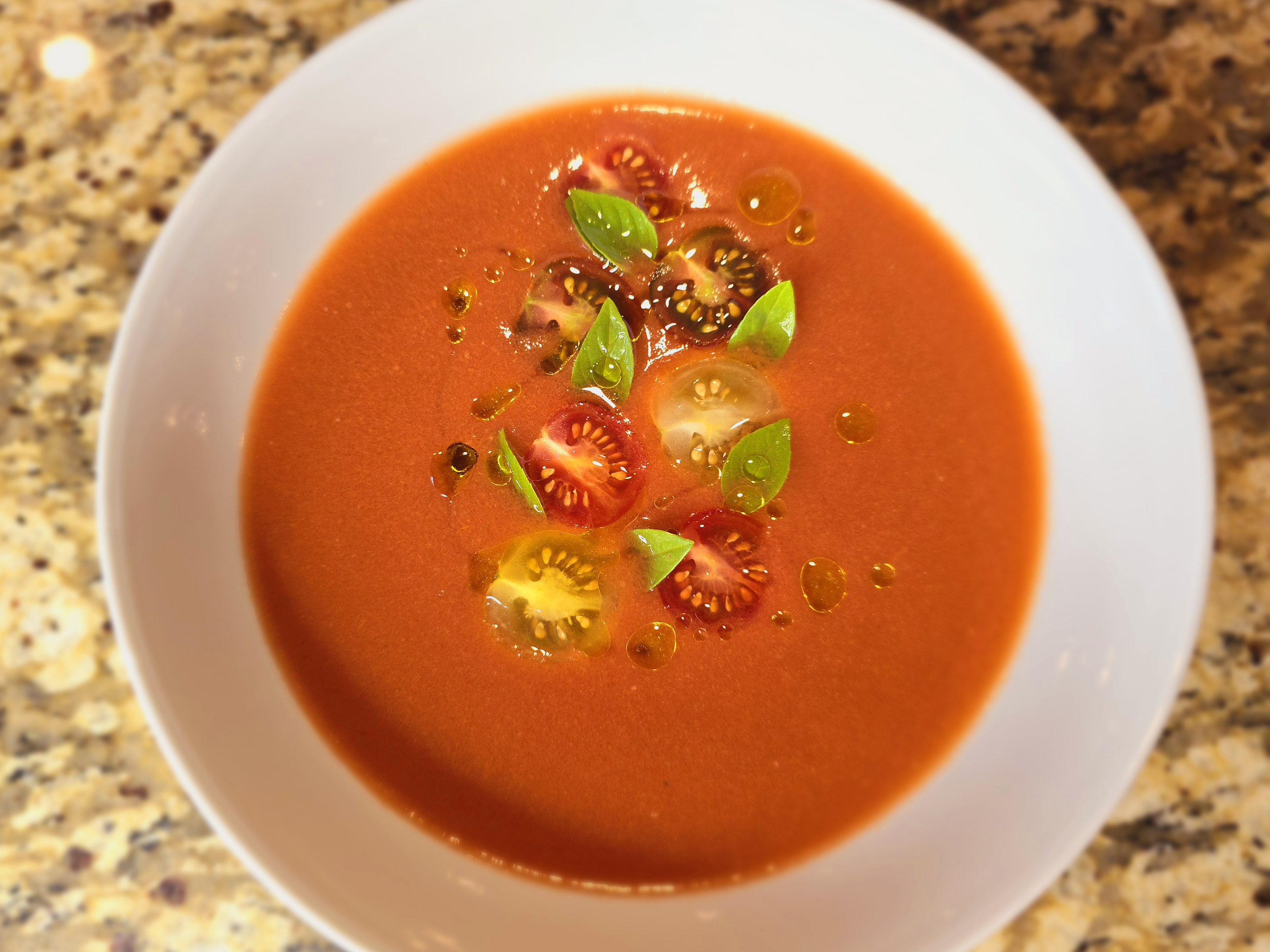 Bowl of vibrant red gazpacho soup garnished with diced vegetables and olive oil drizzle, fresh tomatoes and cucumber visible